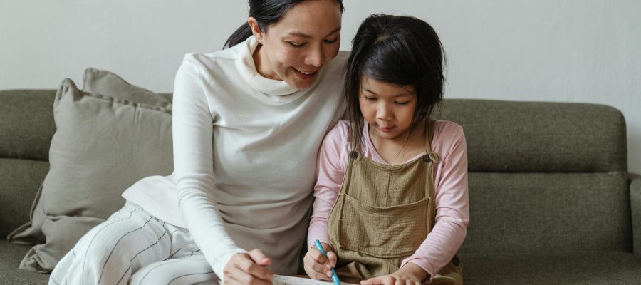 pexels-photo-4473980-4473980 Happy smiling Asian mother and cute little girl sitting on sofa and coloring picture with multicolored crayons