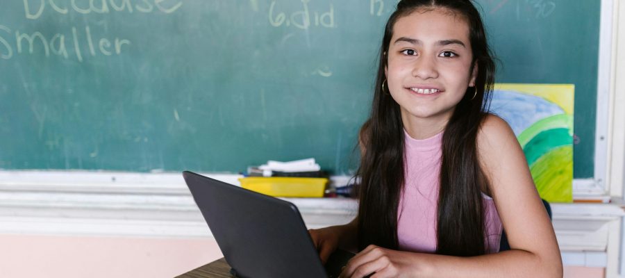 pexels-photo-8500310-8500310 Young girl in a classroom, smiling while using a laptop, against a chalkboard backdrop.